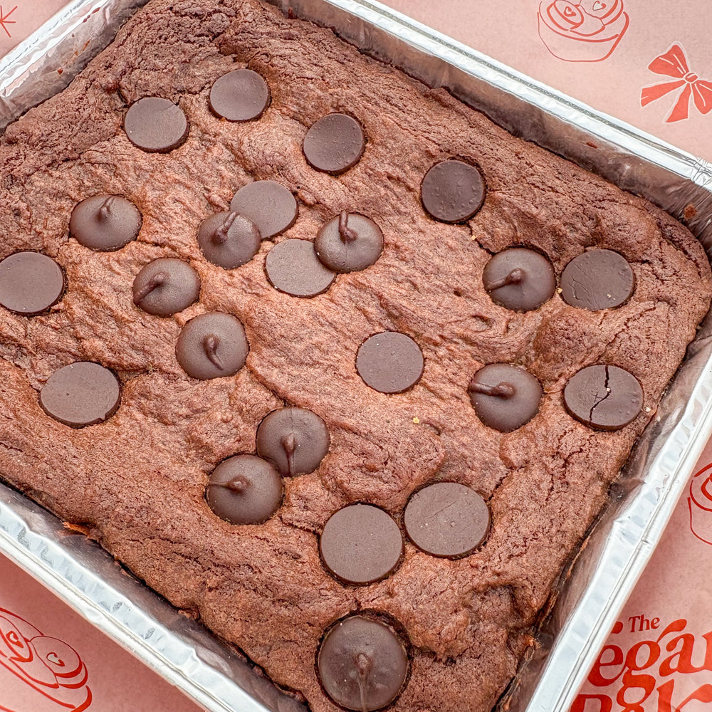 Chocolate cake with chocolate chips in a metal pan on a pink background with 'The Vegan Baker' branding.