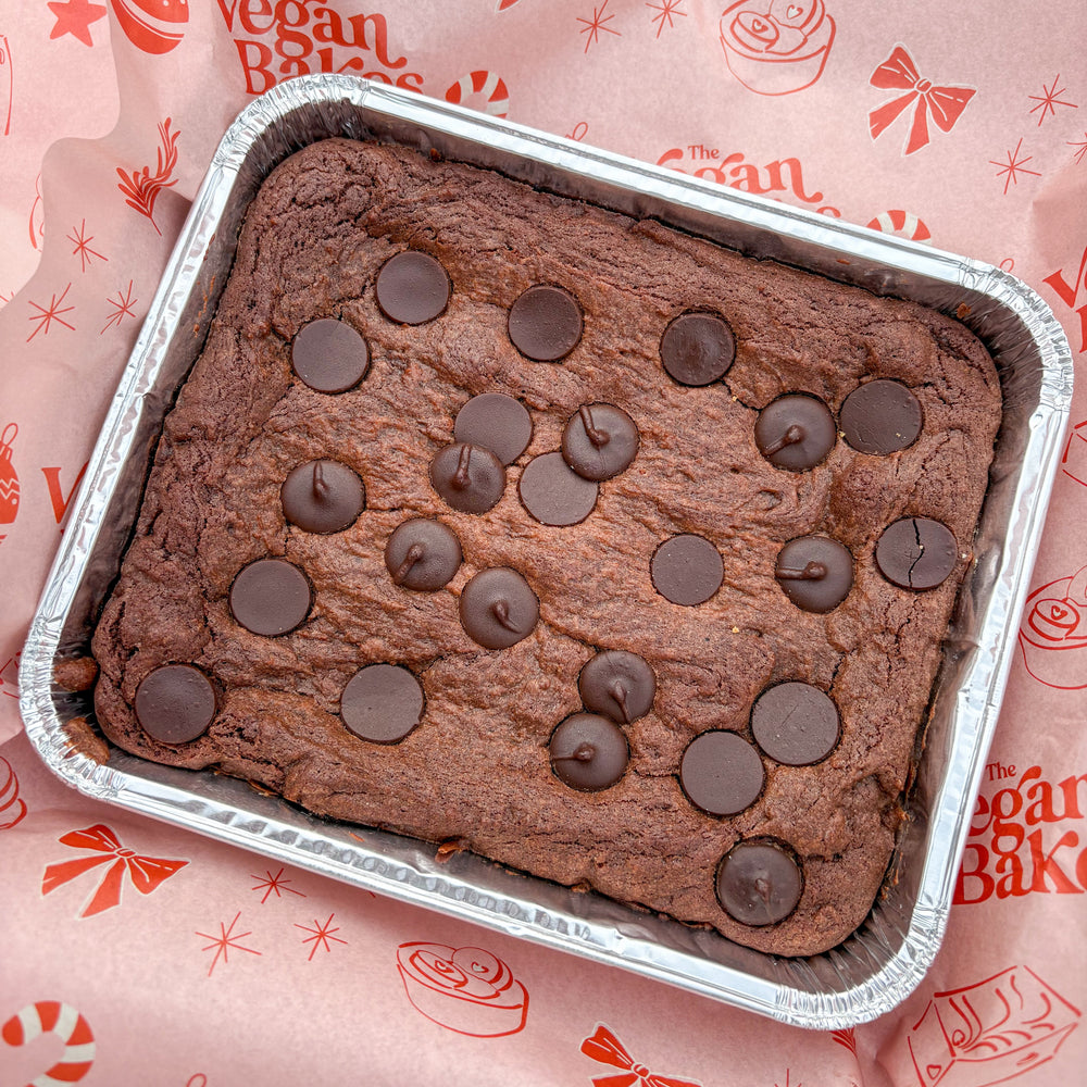 Chocolate cake with choc chips topping in a metal tray on a pink background with 'The Vegan Bake' branding.