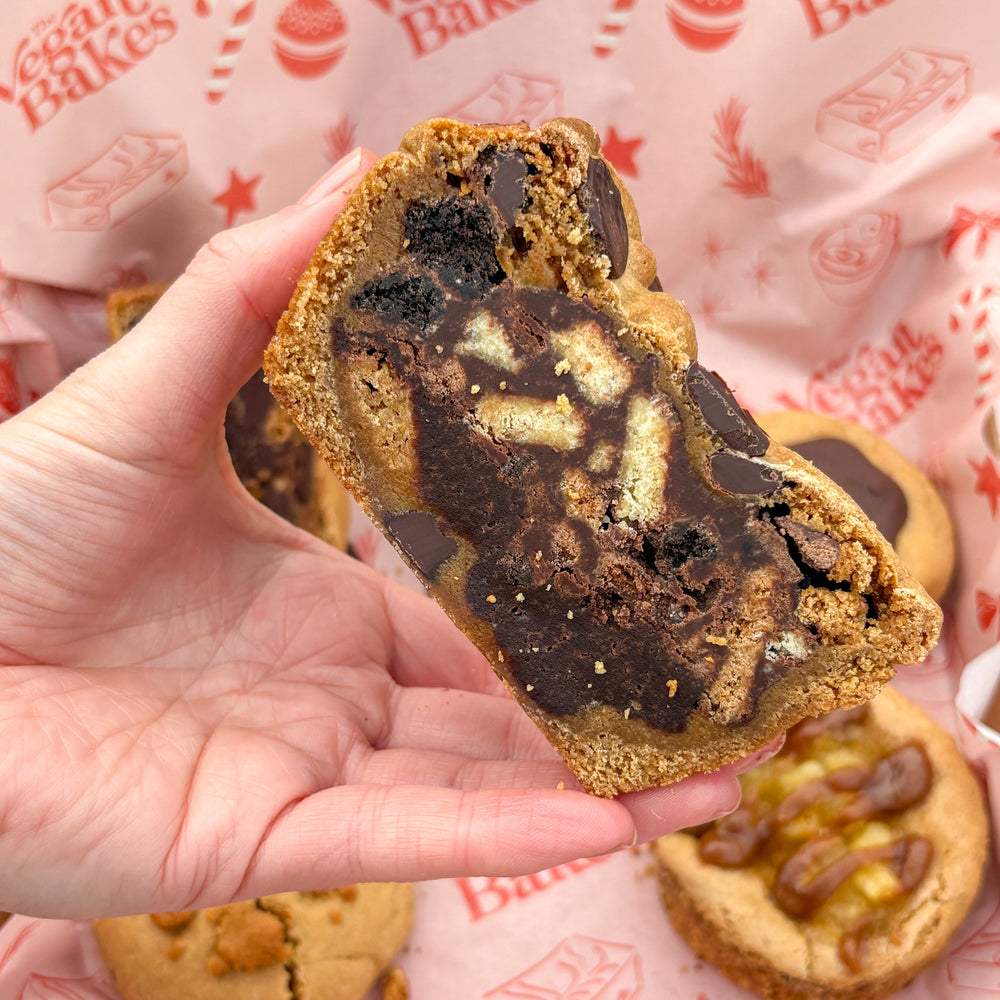 Hand holding a chocolate chip cookie with a visible interior on a 'Vegan Bakes' branded paper background.