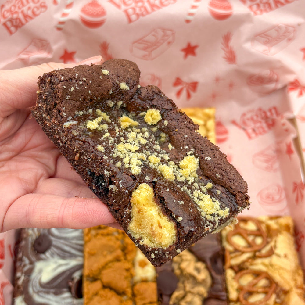 Hand holding a chocolate brownie with a pattern of yellow crumbly topping over a box of assorted brownies.